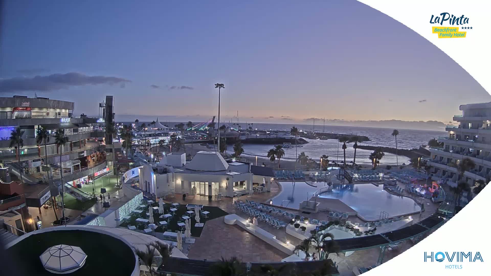 An outdoor evening scene shows a resort with pools and surrounding buildings near a calm ocean under a twilight sky.