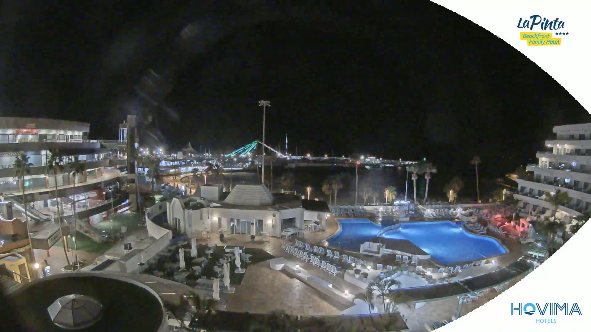 A nighttime aerial view of a resort complex with a swimming pool, situated near a calm body of water and a lit bridge.