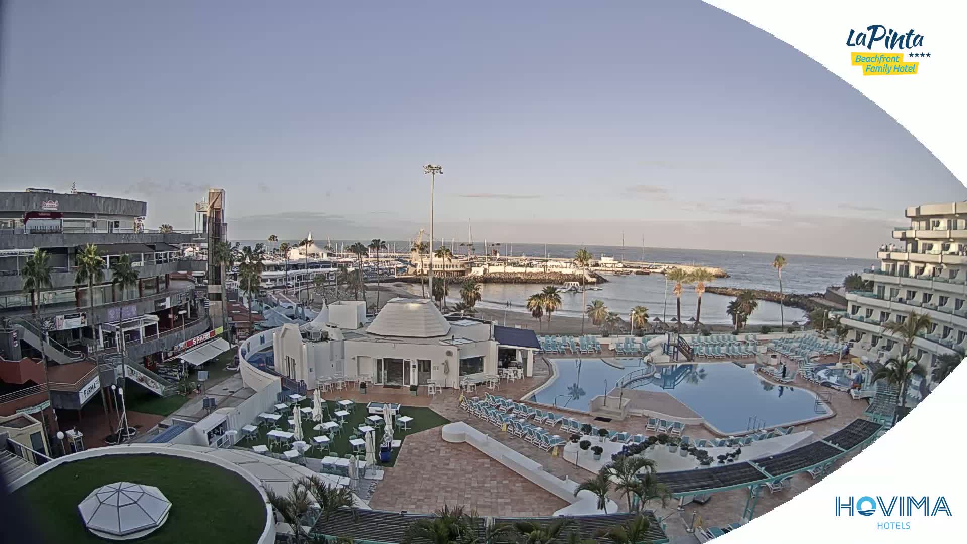 An aerial view shows a resort pool area with a beach and marina beyond under a clear, daytime sky.