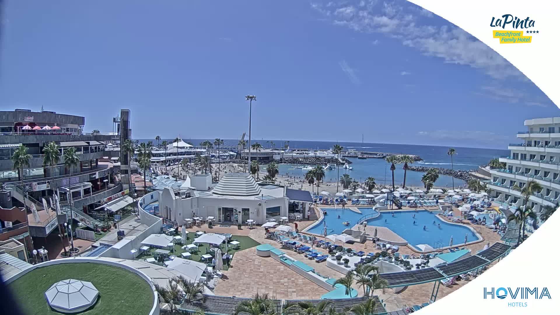 A sunny day reveals a resort pool area, beach, and marina, all situated near a waterfront shopping center.