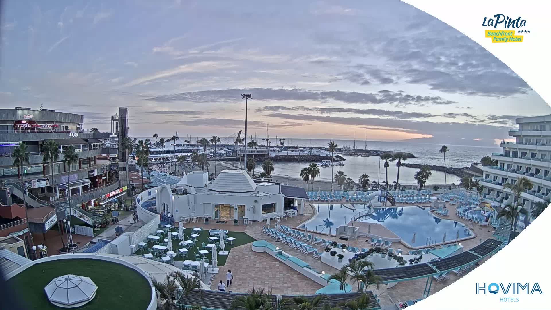 An aerial view of a resort with a swimming pool and beach area at sunset under a partly cloudy sky.