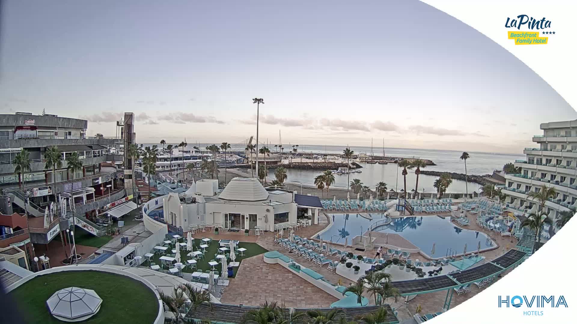 An outdoor scene shows a resort with a swimming pool, beach, marina, and shops under a mostly clear sky.