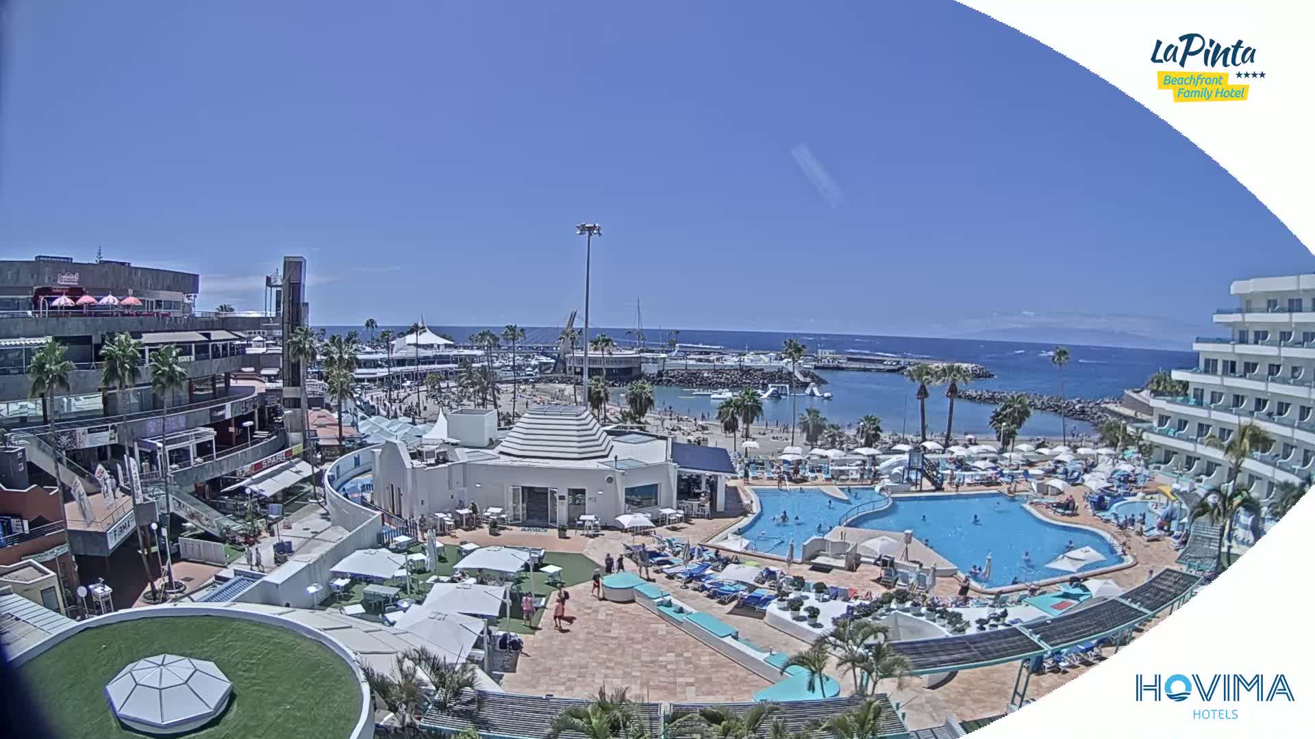 An aerial view of a resort on a sunny day shows a beach, calm ocean, multiple pools, and buildings with palm trees.