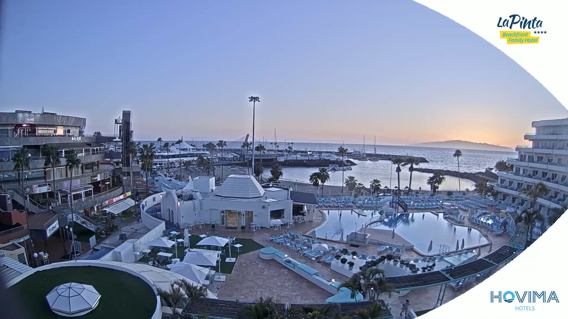 An aerial view of a resort at sunset, showing a swimming pool, beach, marina, and buildings under a clear sky.