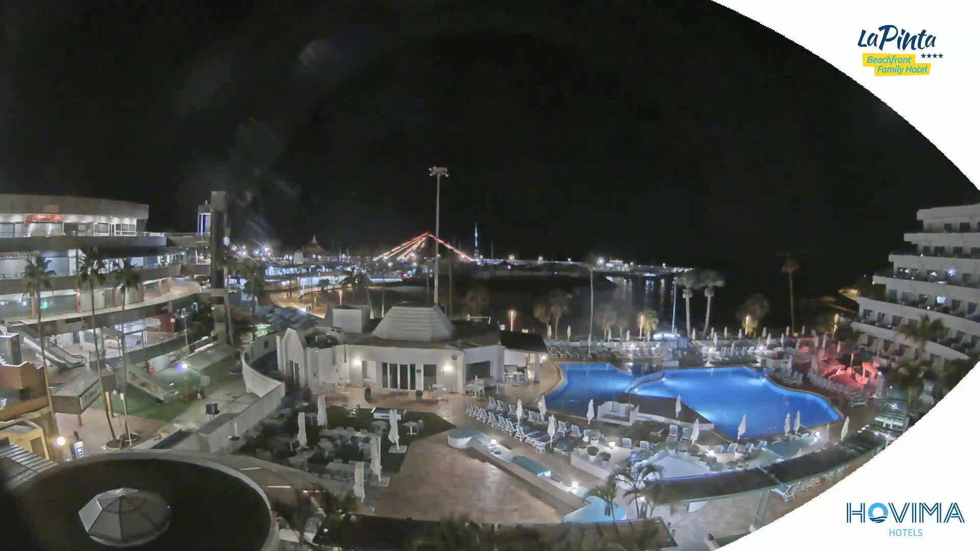 A nighttime aerial view of a resort complex with swimming pools, surrounding buildings, and a harbor in the distance under a clear, dark sky.