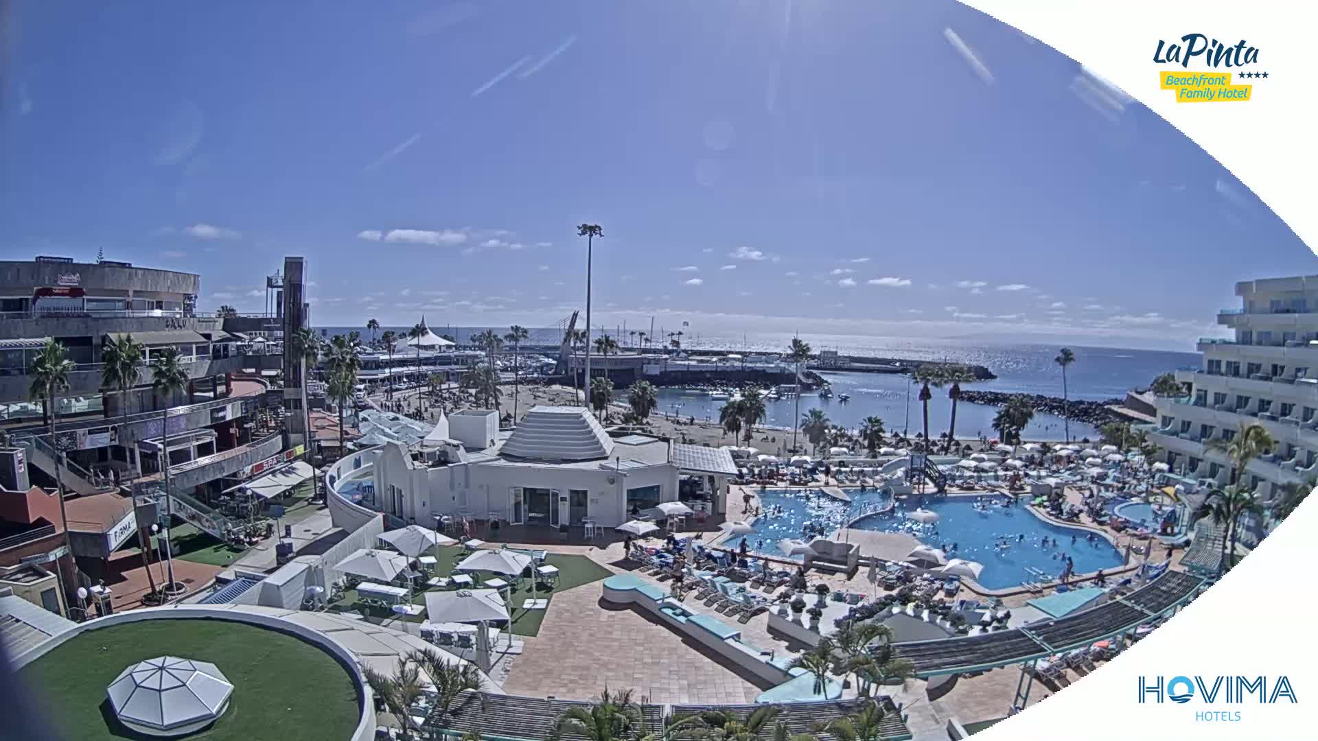 An aerial view shows a resort with multiple pools and a beach under sunny skies, next to a marina and shops.