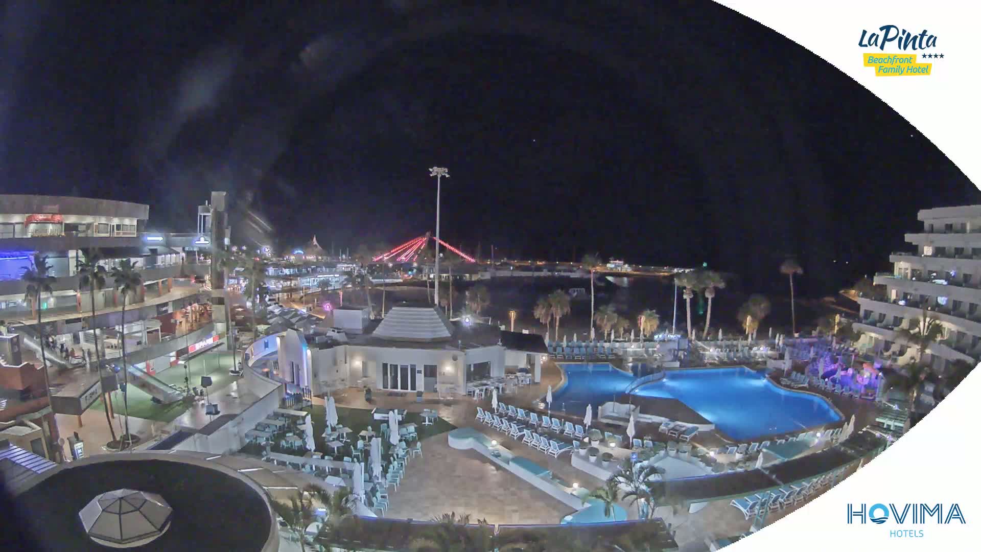 A nighttime aerial view of a resort with swimming pools, palm trees, and a brightly lit waterfront area in the background under a clear, dark sky.