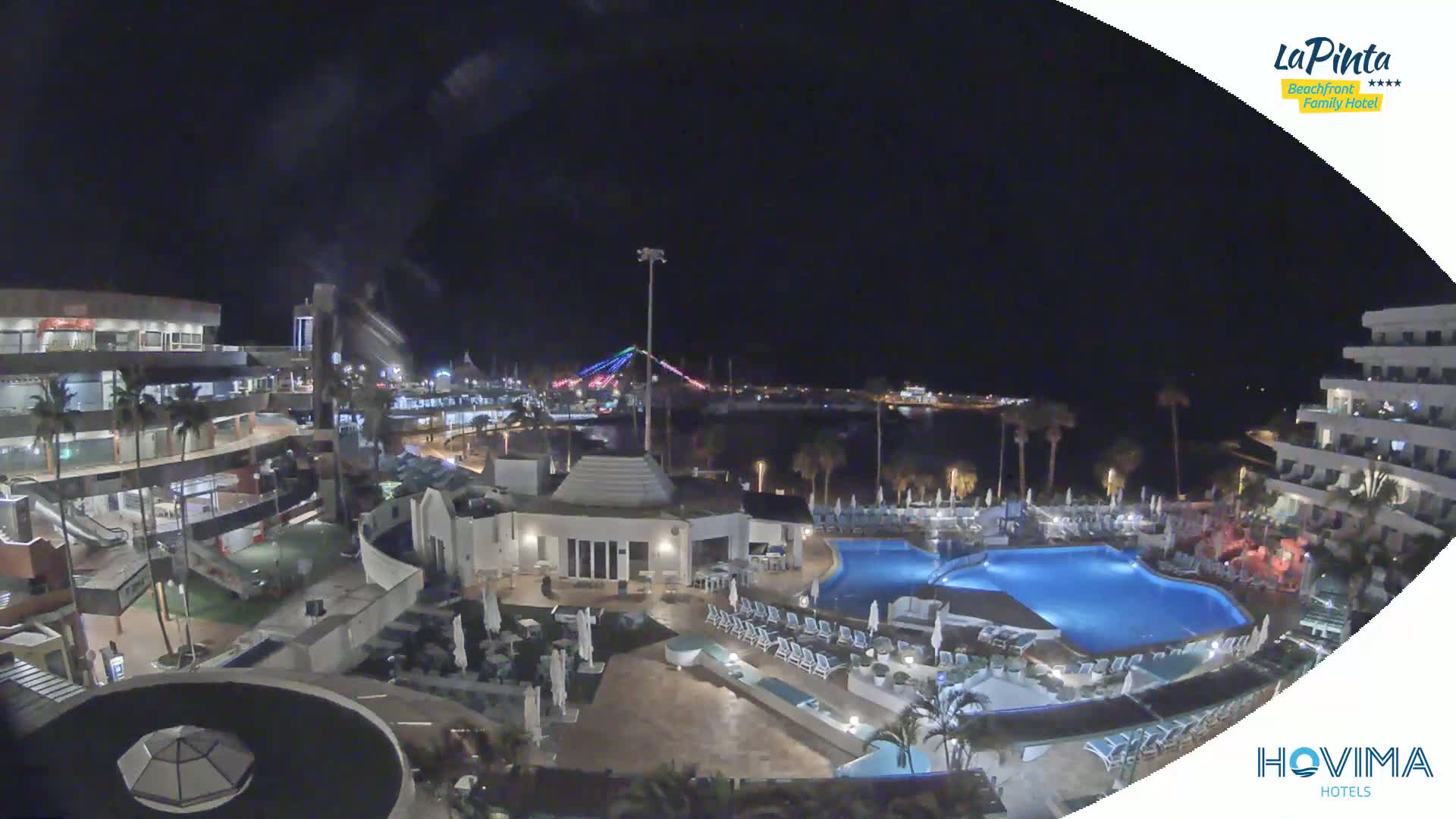 A nighttime aerial view shows a resort complex with a swimming pool, palm trees, and a body of water in the background, illuminated by lights.