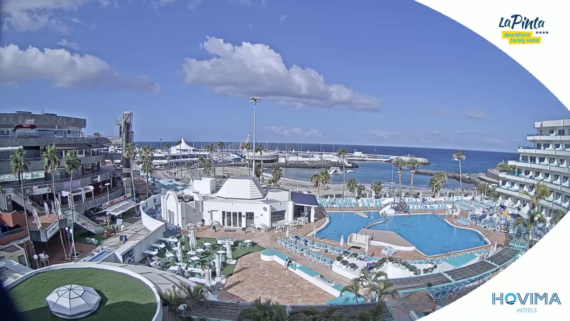 A resort with a swimming pool and surrounding buildings overlooks a marina and sandy beach under a partly cloudy sky.