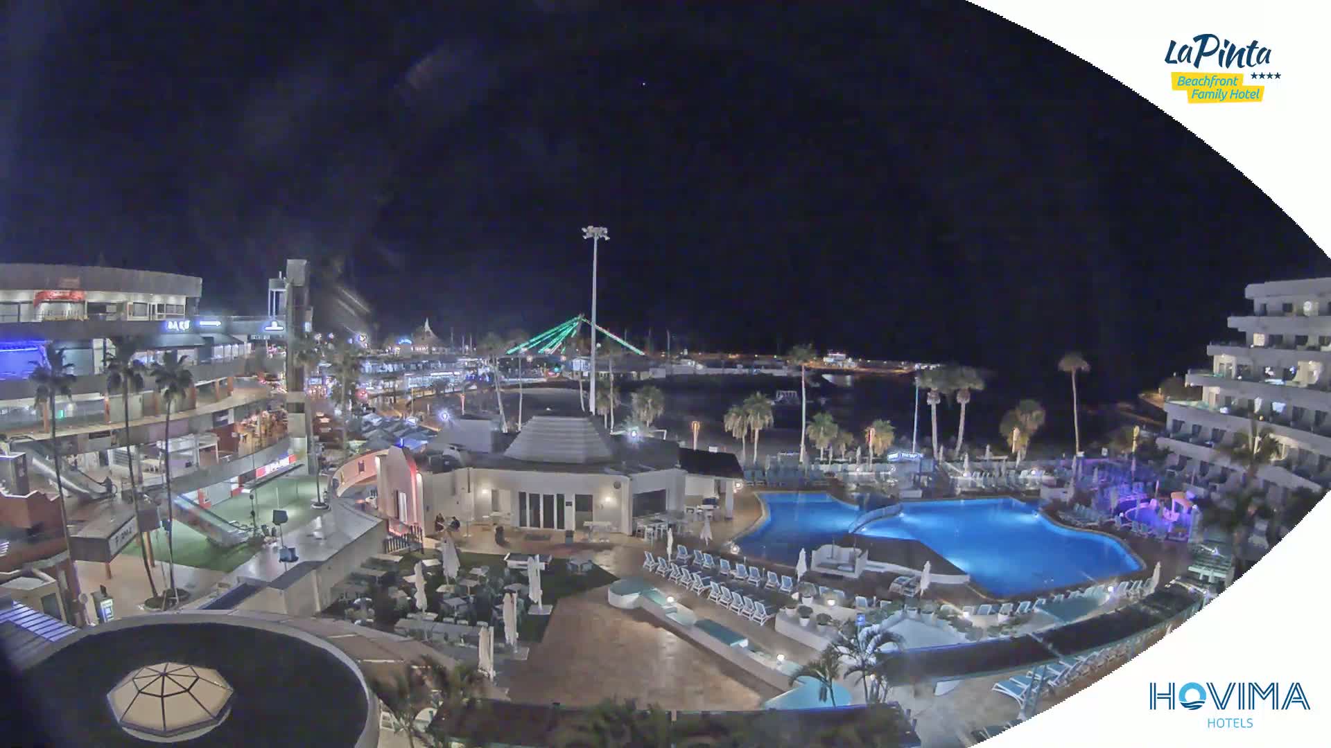 A nighttime aerial view shows a resort complex with swimming pools, palm trees, and a beachfront area illuminated under a dark sky.