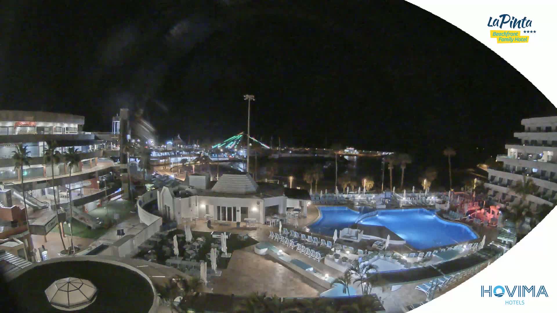 A nighttime aerial view of a resort complex with a swimming pool, surrounding buildings, and a waterfront in the distance.