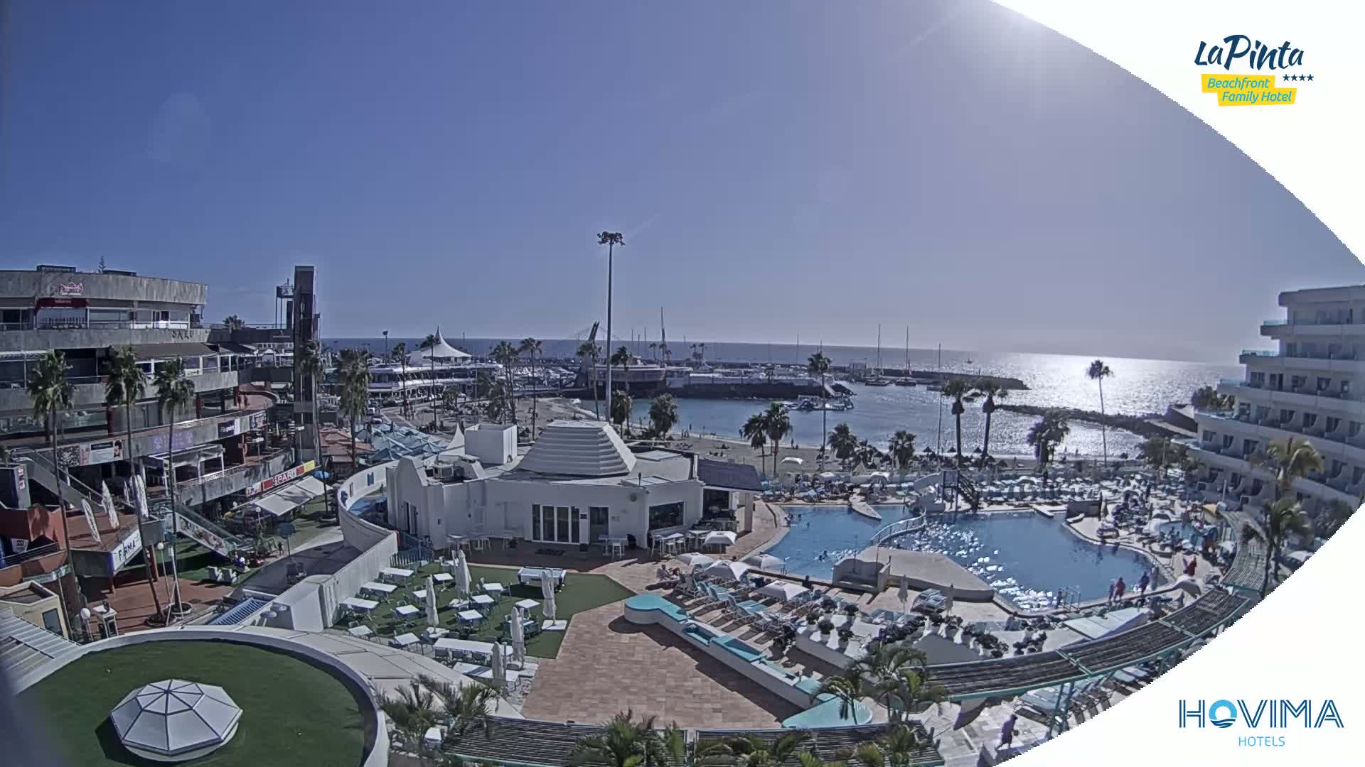 An aerial view shows a resort complex with pools and a marina on a sunny day, overlooking a calm ocean.