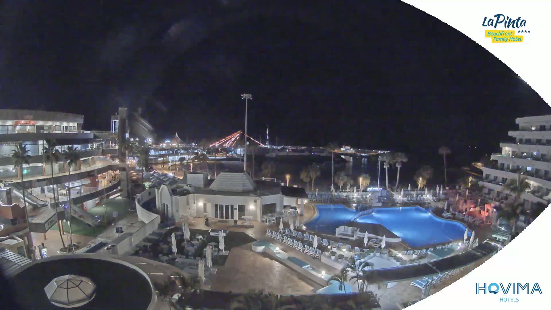 A nighttime aerial view of a resort complex with swimming pools, surrounded by buildings and palm trees, overlooks a body of water with a bridge in the distance under a clear, dark sky.