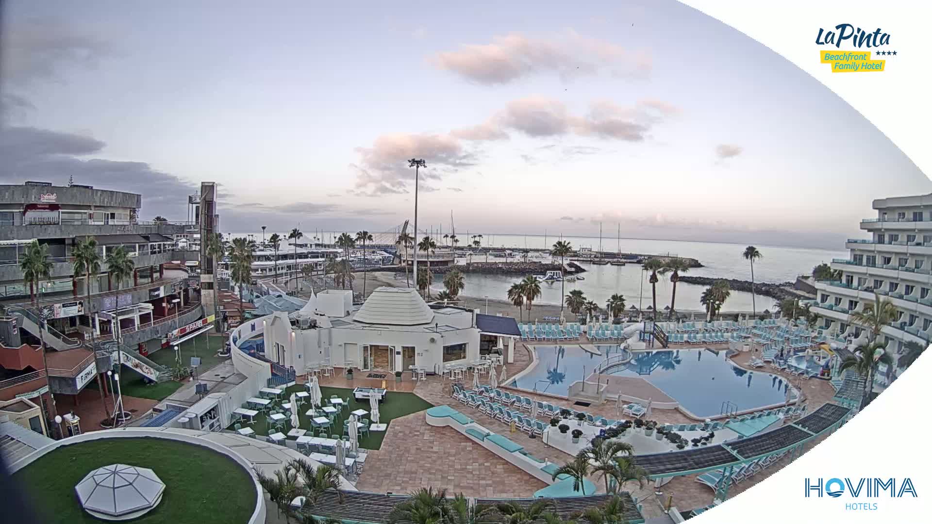 An aerial view shows a resort complex with multiple pools, a marina, and buildings under a mostly clear, pale sky at dawn or dusk.