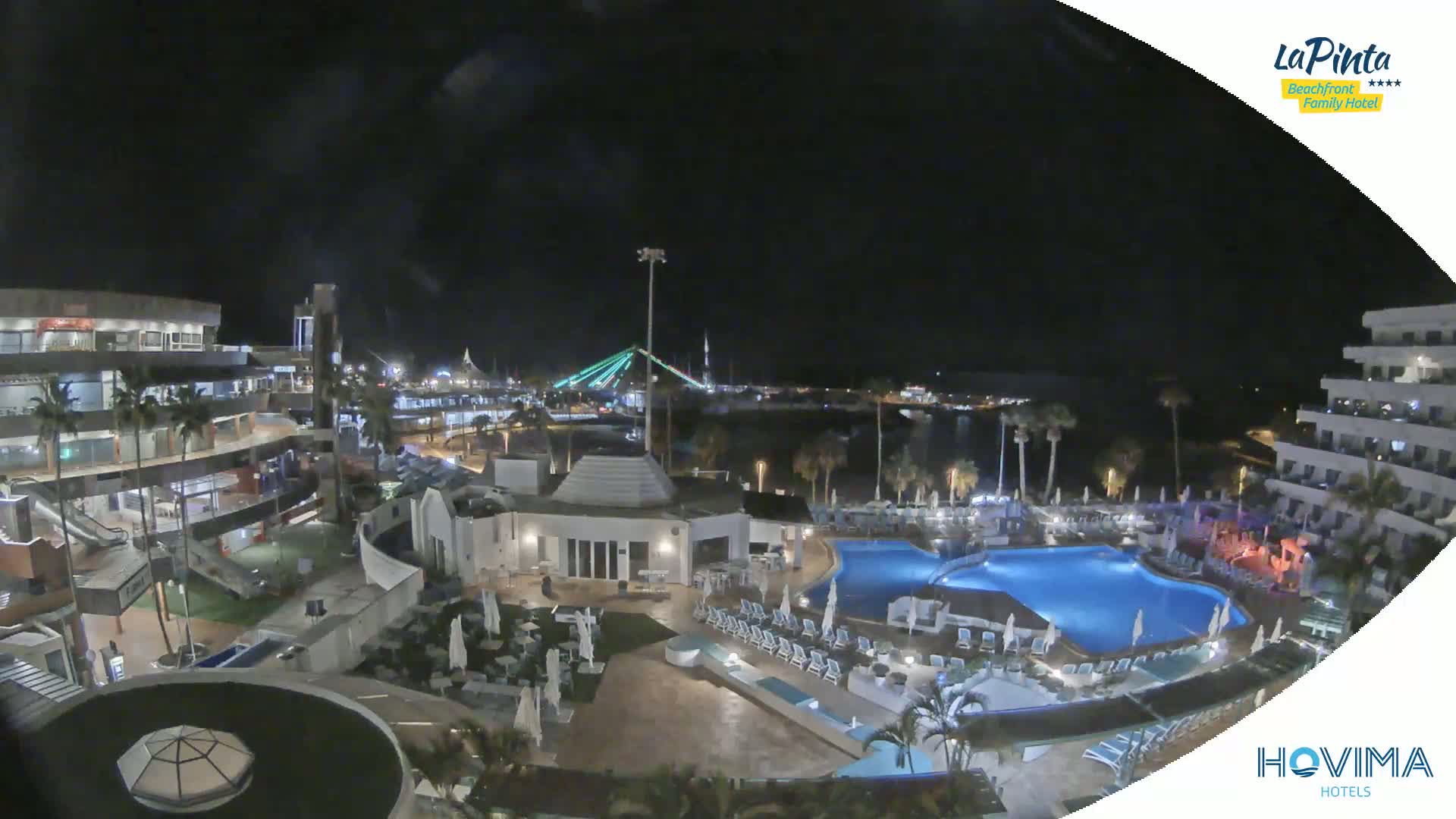 A nighttime aerial view of a resort complex with pools, surrounding buildings, and a lit bridge in the distance.
