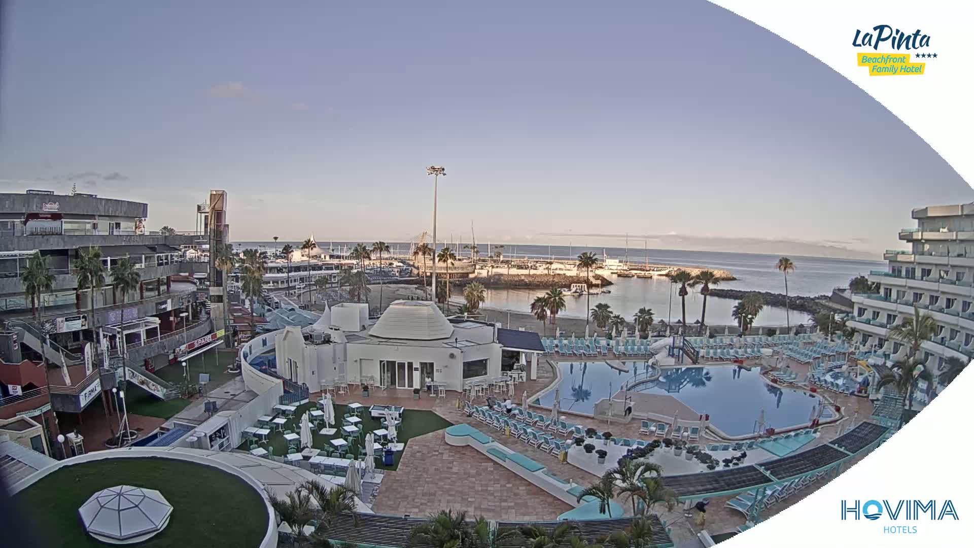 A high-angle, daytime view shows a resort pool area adjacent to a marina and calm ocean under a clear sky.