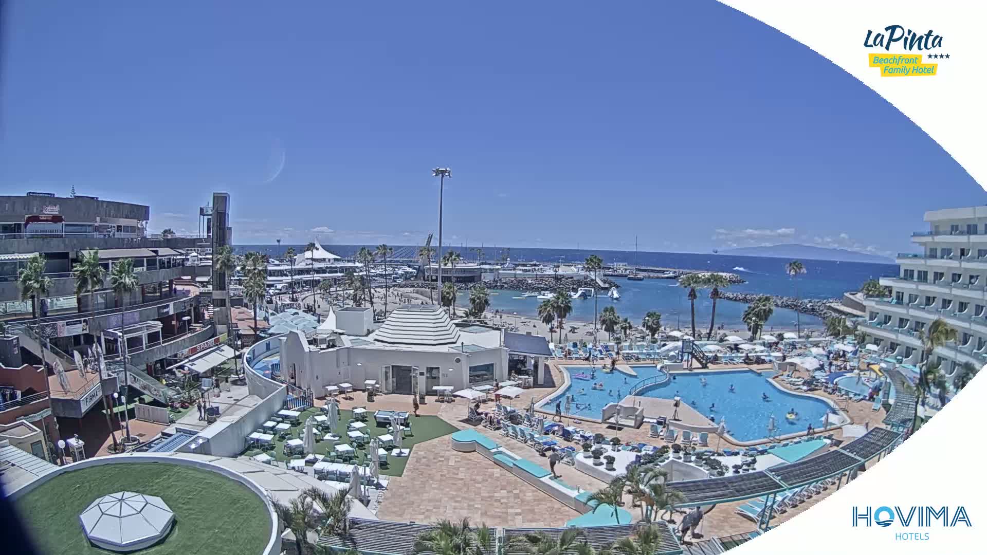 An aerial view shows a resort with a swimming pool complex next to a beach and marina on a sunny day.