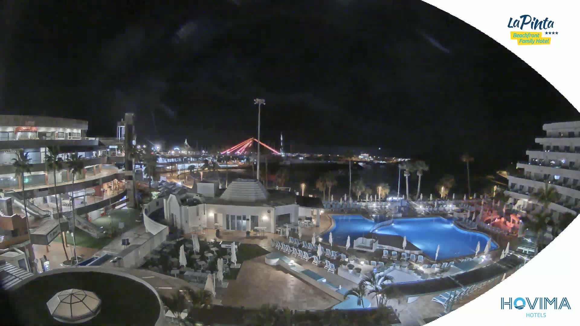 A nighttime aerial view of a resort complex with multiple pools and buildings, overlooking a body of water and a lit bridge under a clear, dark sky.