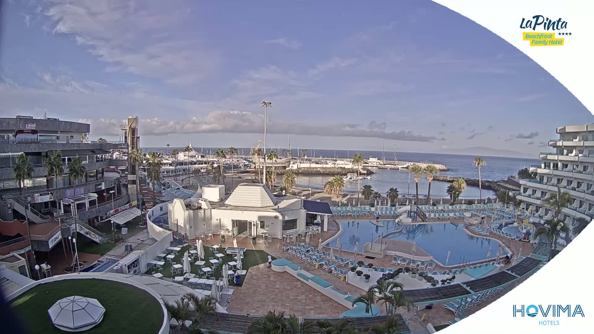 A resort with a swimming pool overlooks a marina filled with boats on a partly cloudy day.