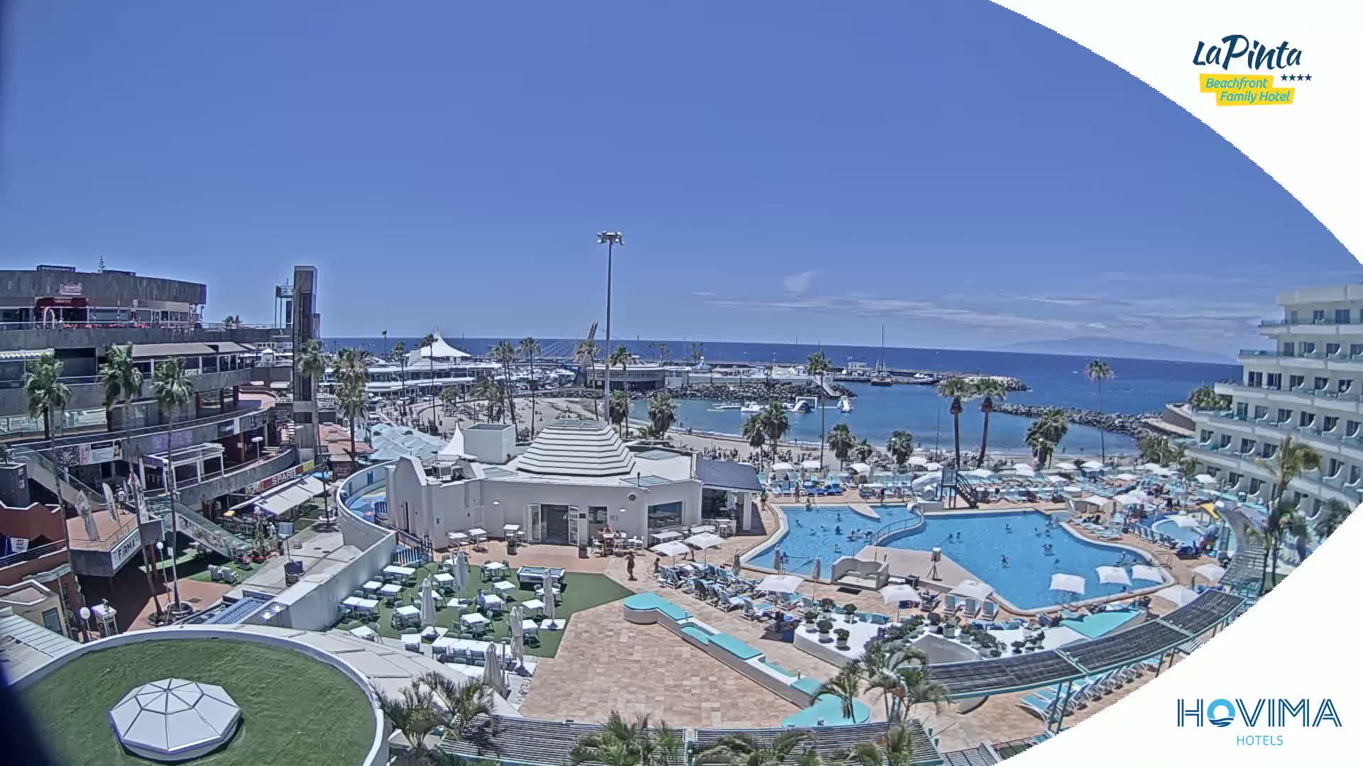 A resort with multiple pools is situated next to a harbor and beach on a sunny day.