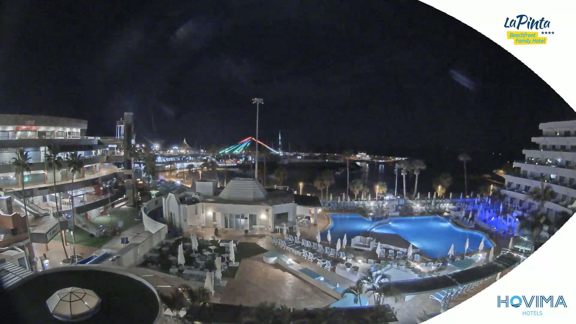 A nighttime aerial view of a resort complex with swimming pools, surrounding buildings, and a brightly lit bridge in the distance.