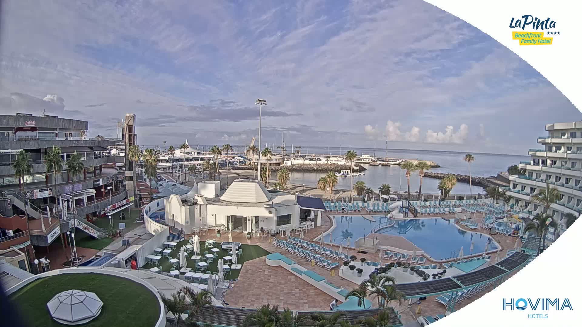A partly cloudy day reveals a resort pool area, adjacent shops and restaurants, and a marina filled with boats.