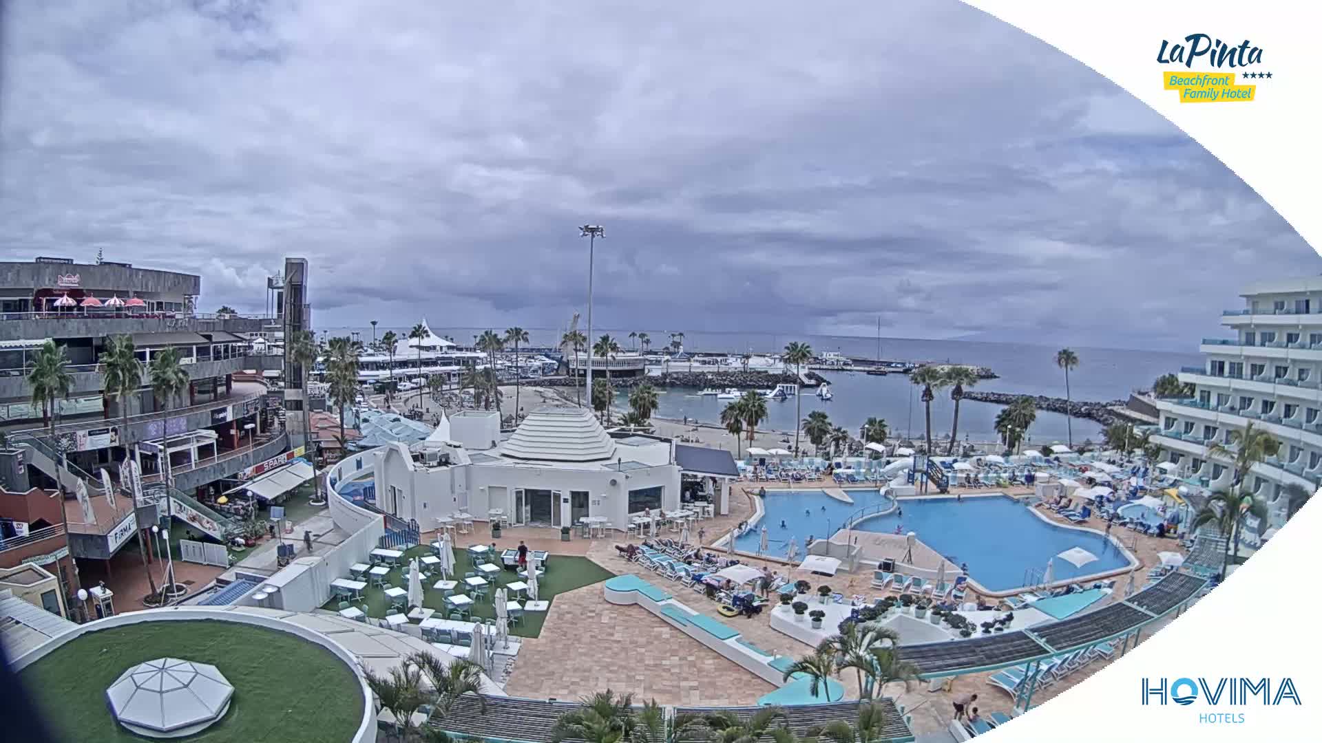 An overcast day reveals a resort with a pool and surrounding buildings overlooking a calm harbor and ocean.