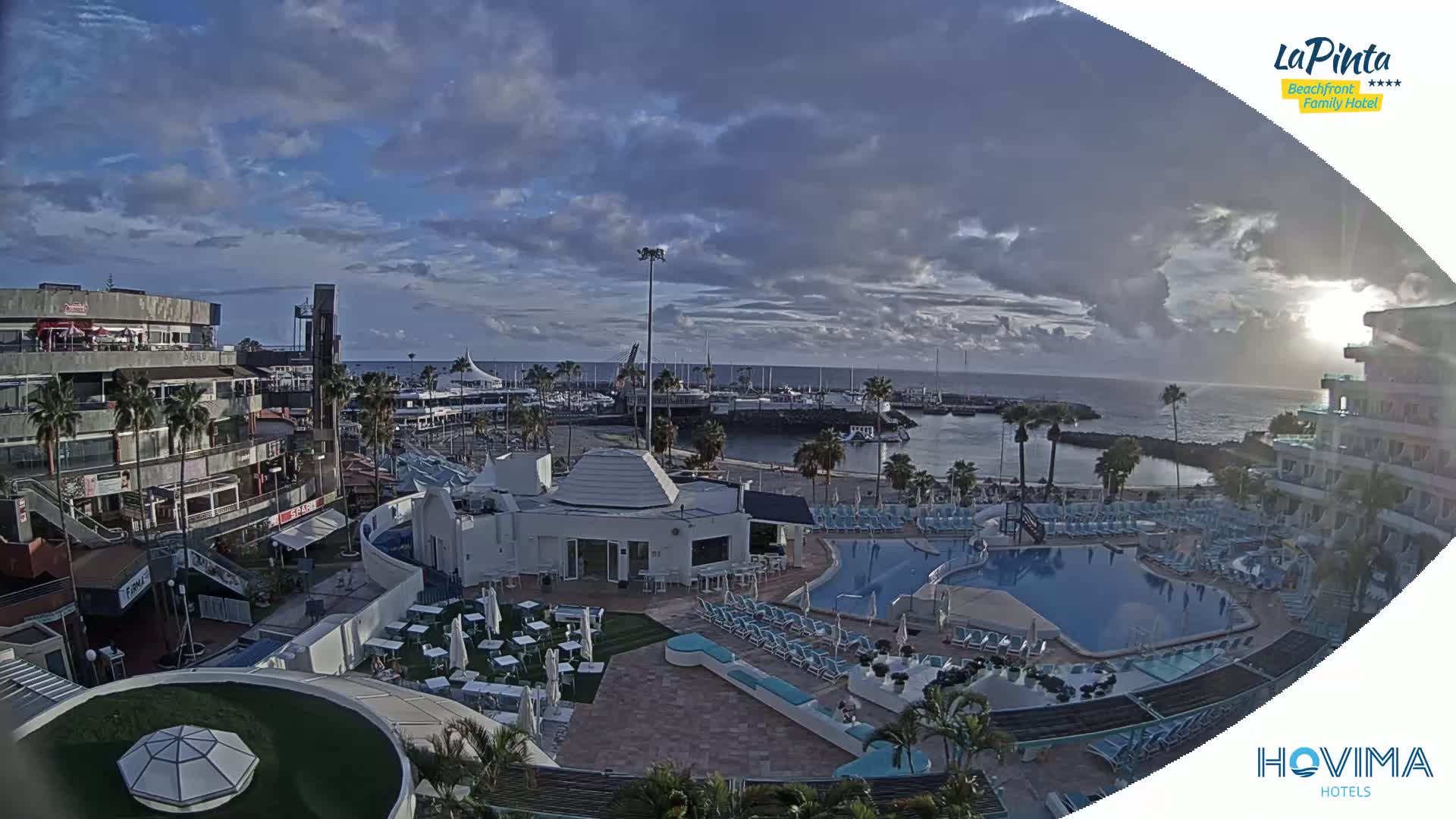 A partly cloudy sunrise over a resort town with a marina, featuring a swimming pool and surrounding buildings.