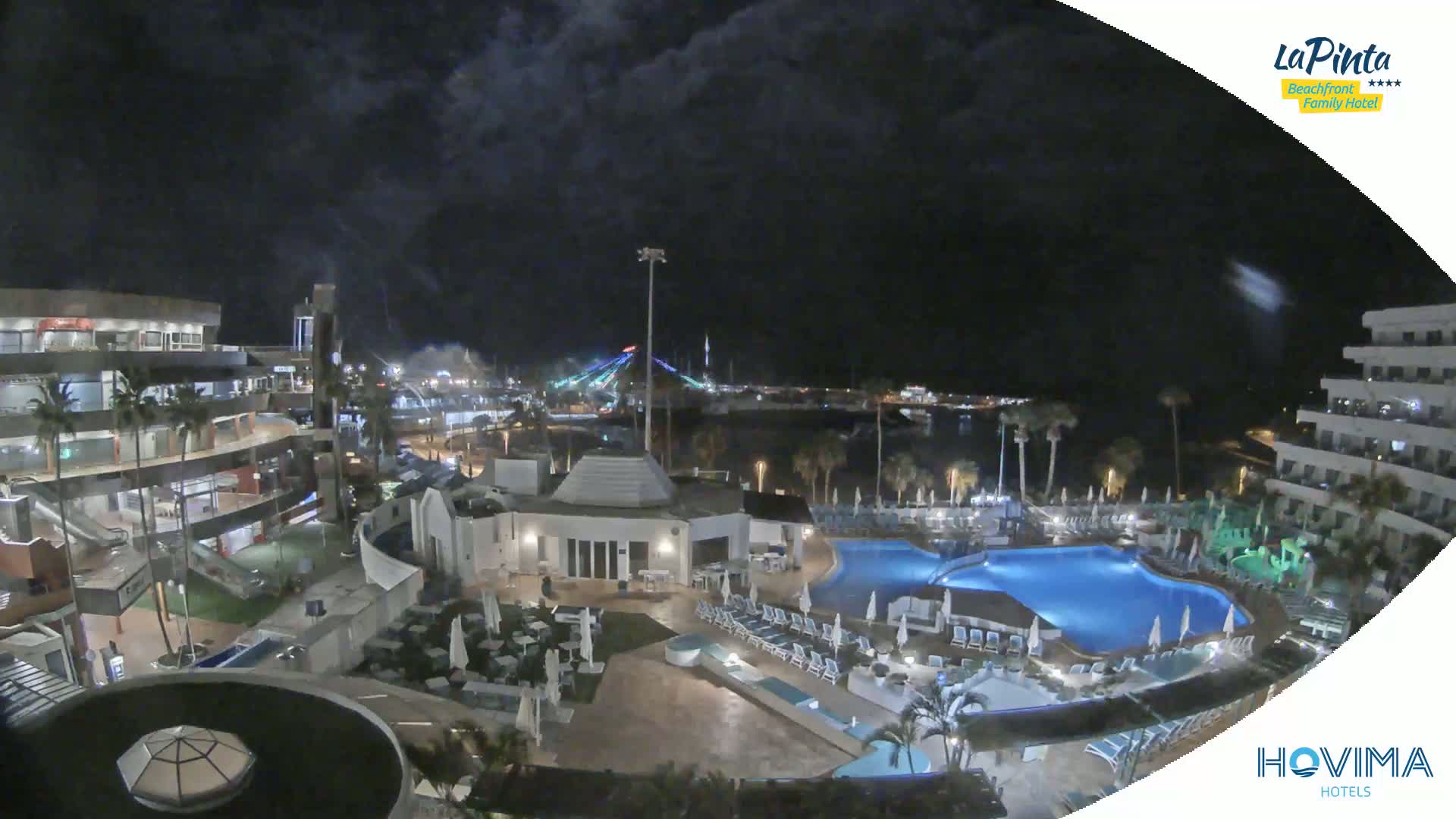 A nighttime aerial view shows a resort with swimming pools, palm trees, and buildings illuminated against a dark, cloudy sky.