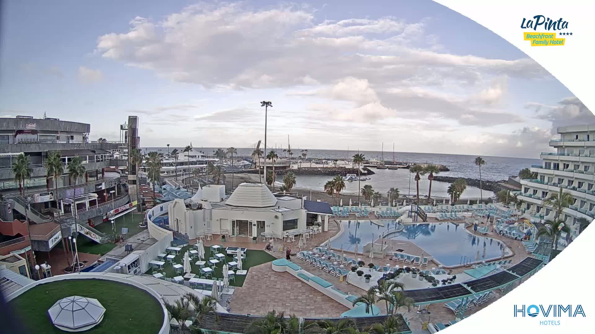 An outdoor scene shows a resort with a swimming pool, surrounding buildings, and a small harbor with boats under a partly cloudy sky.