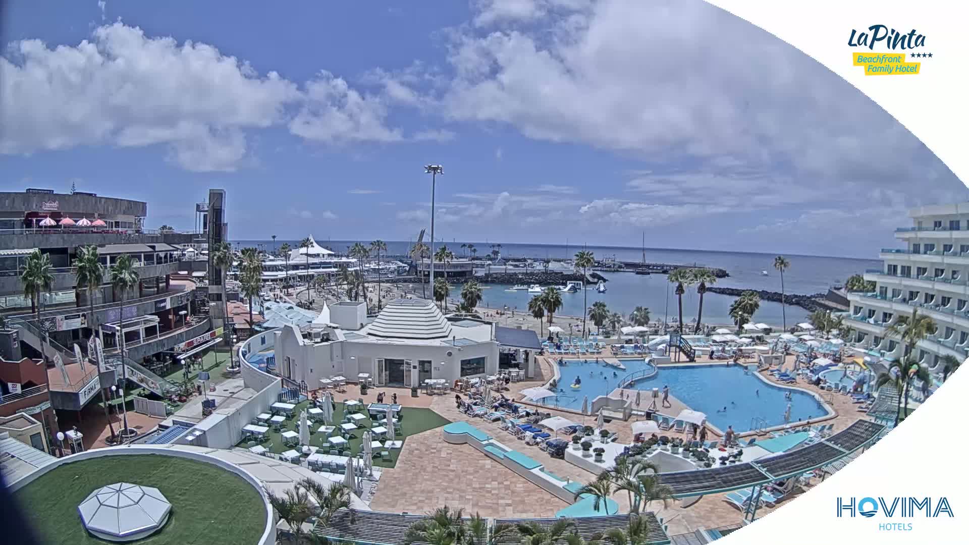 A sunny day overlooks a resort with a pool and beach, a marina, and other buildings.