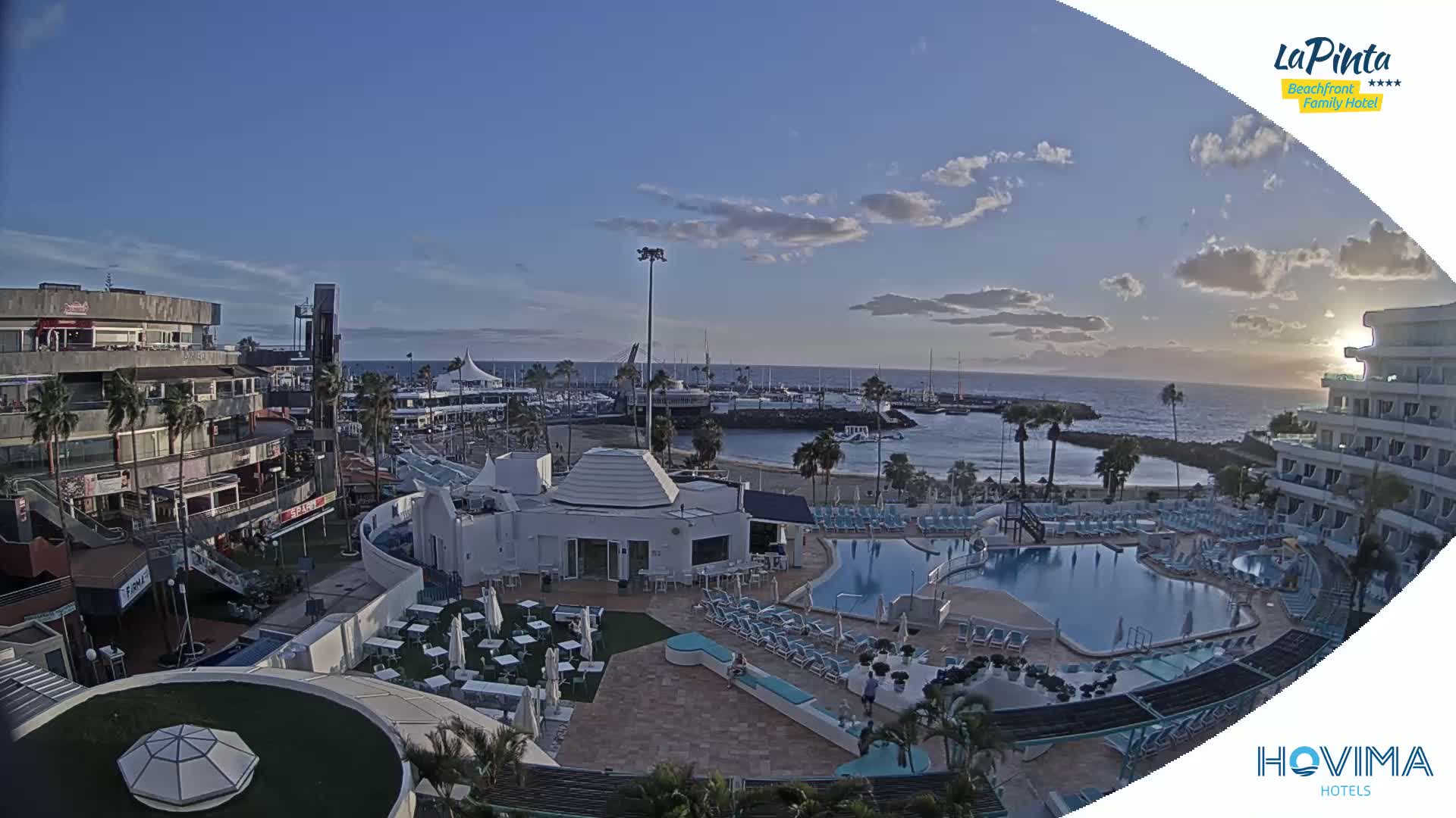 An aerial view of a resort complex at sunset, featuring a swimming pool, beach, marina, and nearby buildings under a partly cloudy sky.