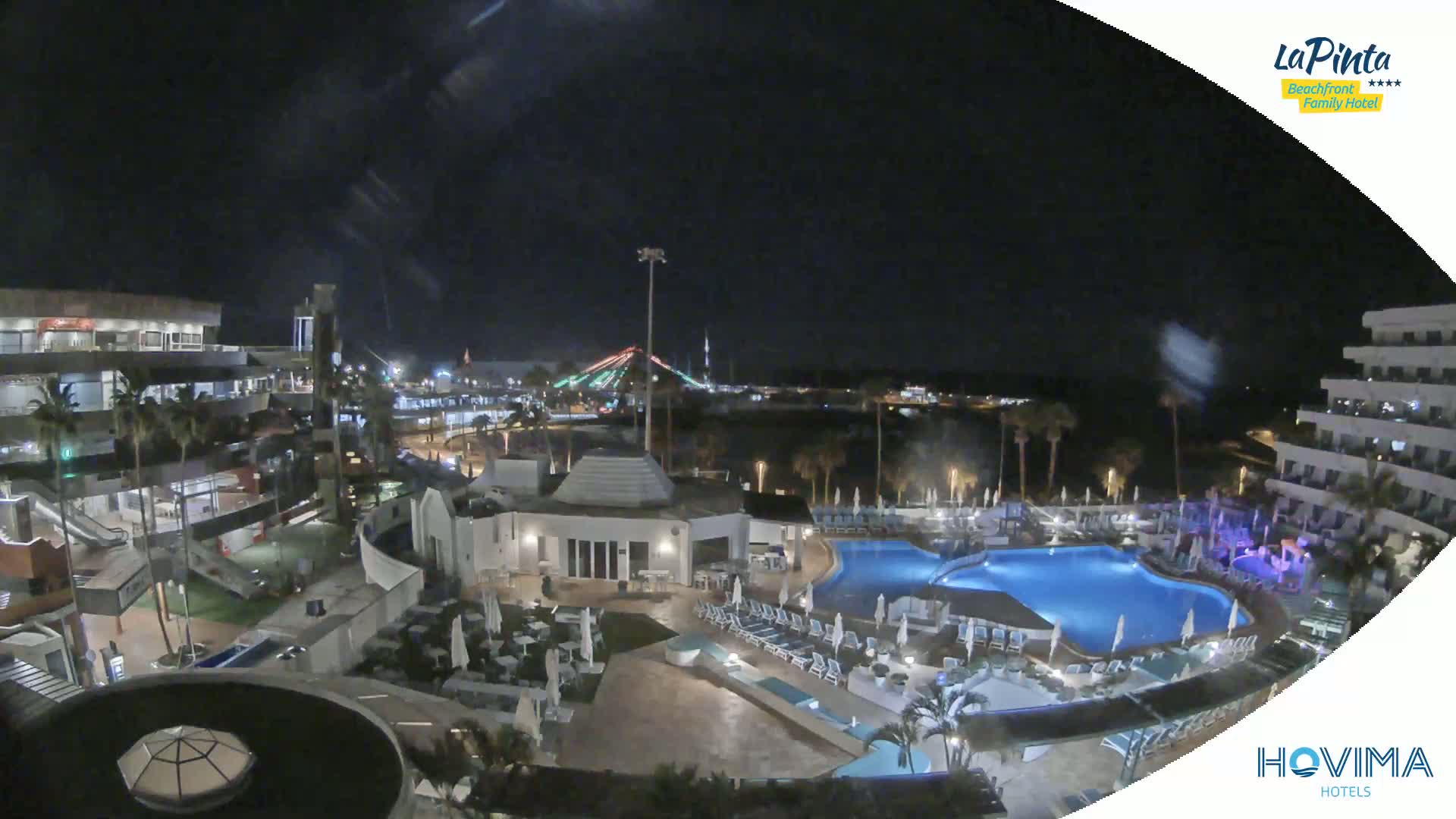 A nighttime aerial view shows a resort complex with a swimming pool, surrounding buildings, and palm trees, under a clear, dark sky.
