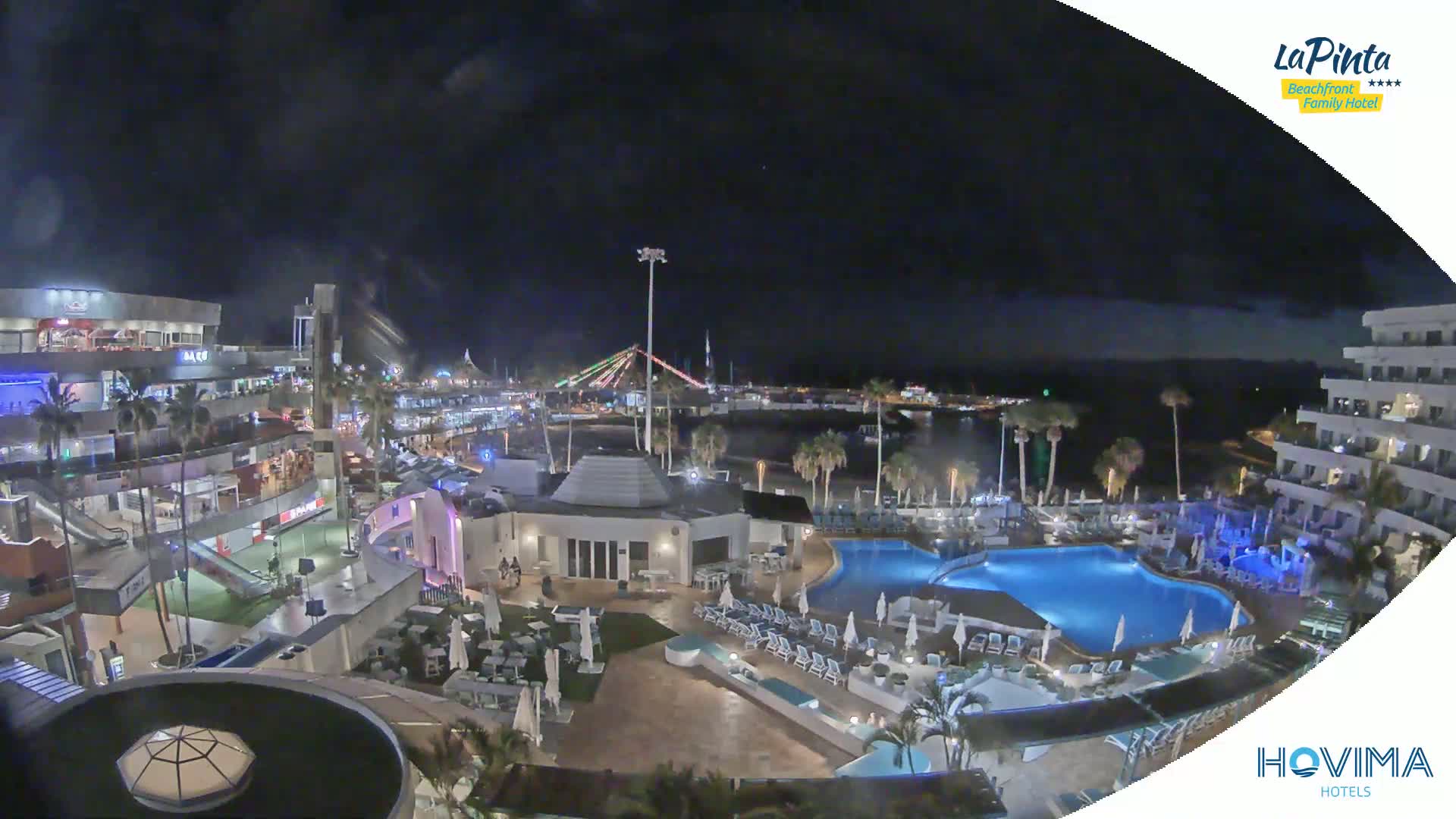 A nighttime aerial view of a resort complex with a swimming pool, surrounded by buildings and palm trees, overlooking a calm body of water under a dark, slightly cloudy sky.