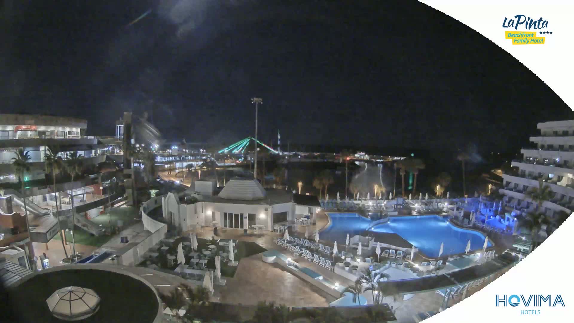 A nighttime aerial view of a resort complex with a swimming pool, surrounding buildings, and a bridge in the distance under a clear, dark sky.