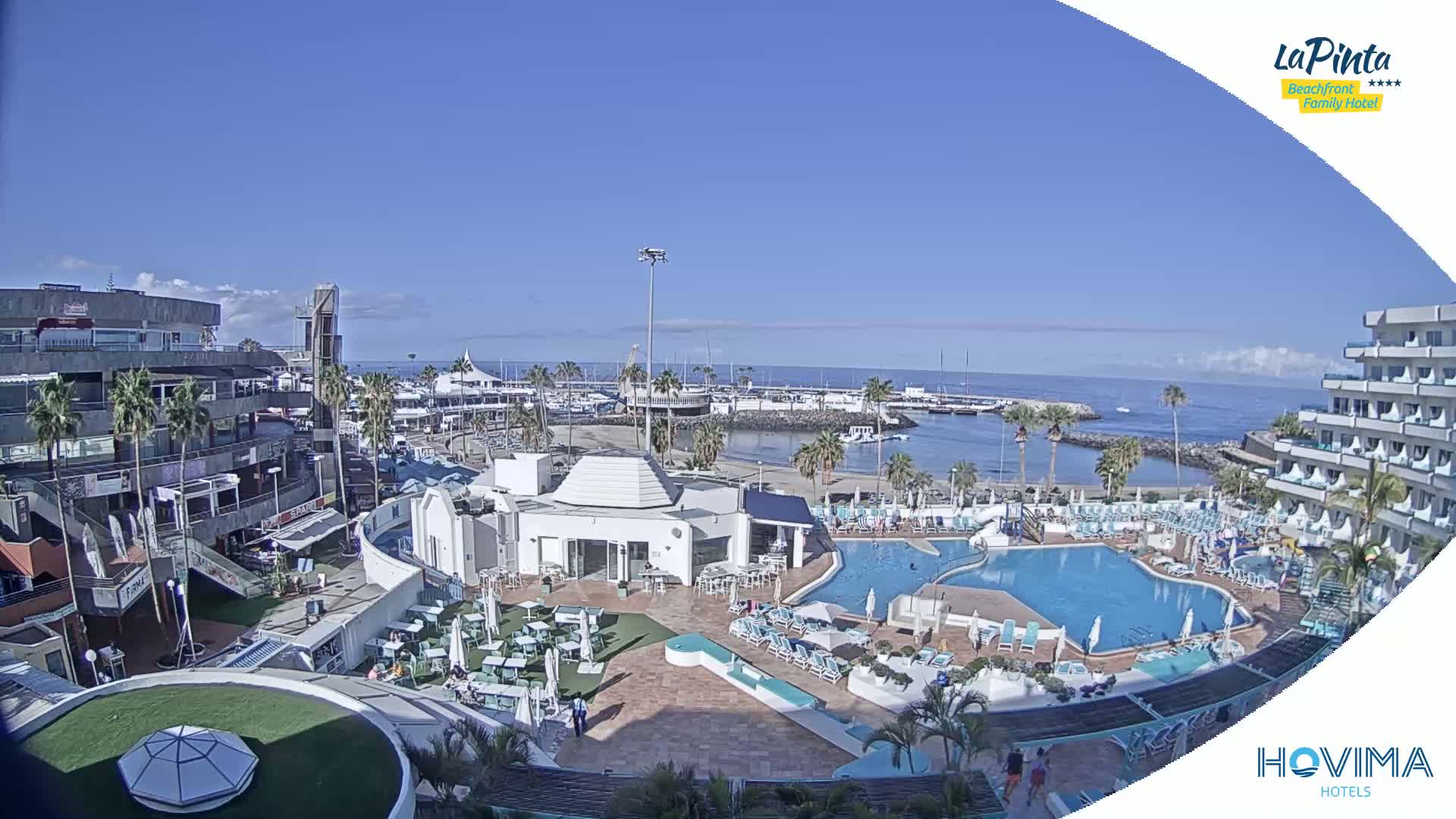 An aerial view shows a resort with a swimming pool and surrounding buildings next to a marina on a sunny day.