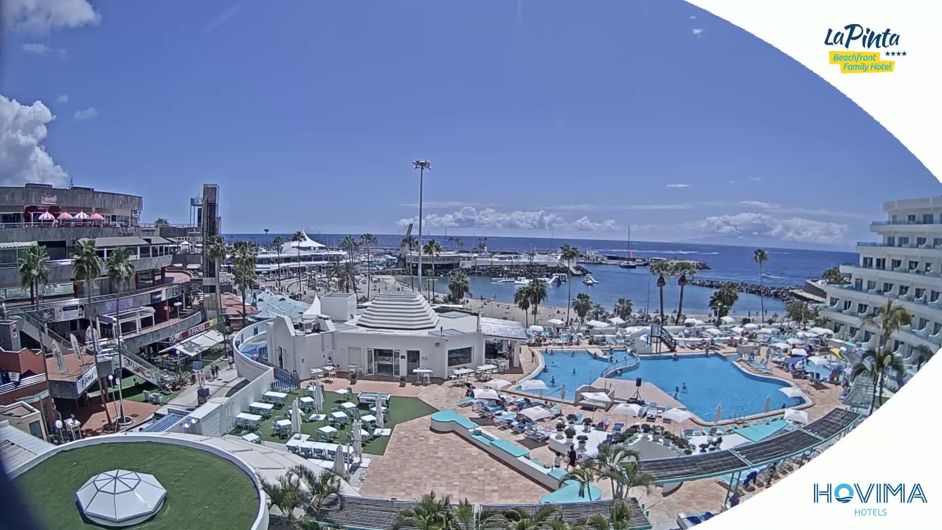 A resort pool area with a beach and marina in view on a sunny day with partly cloudy skies.