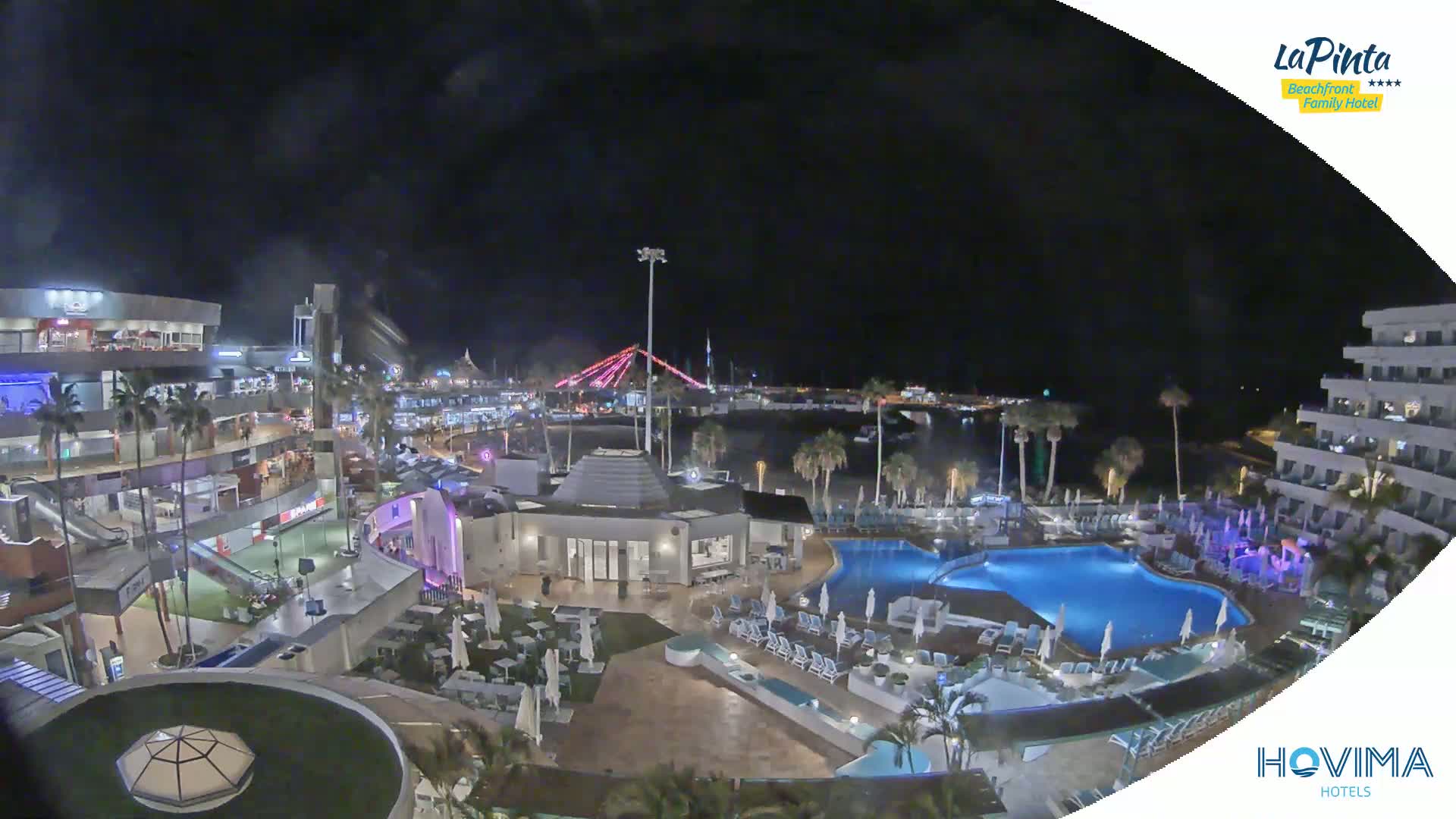A nighttime aerial view shows a resort complex with pools, palm trees, and buildings illuminated against a dark sky.