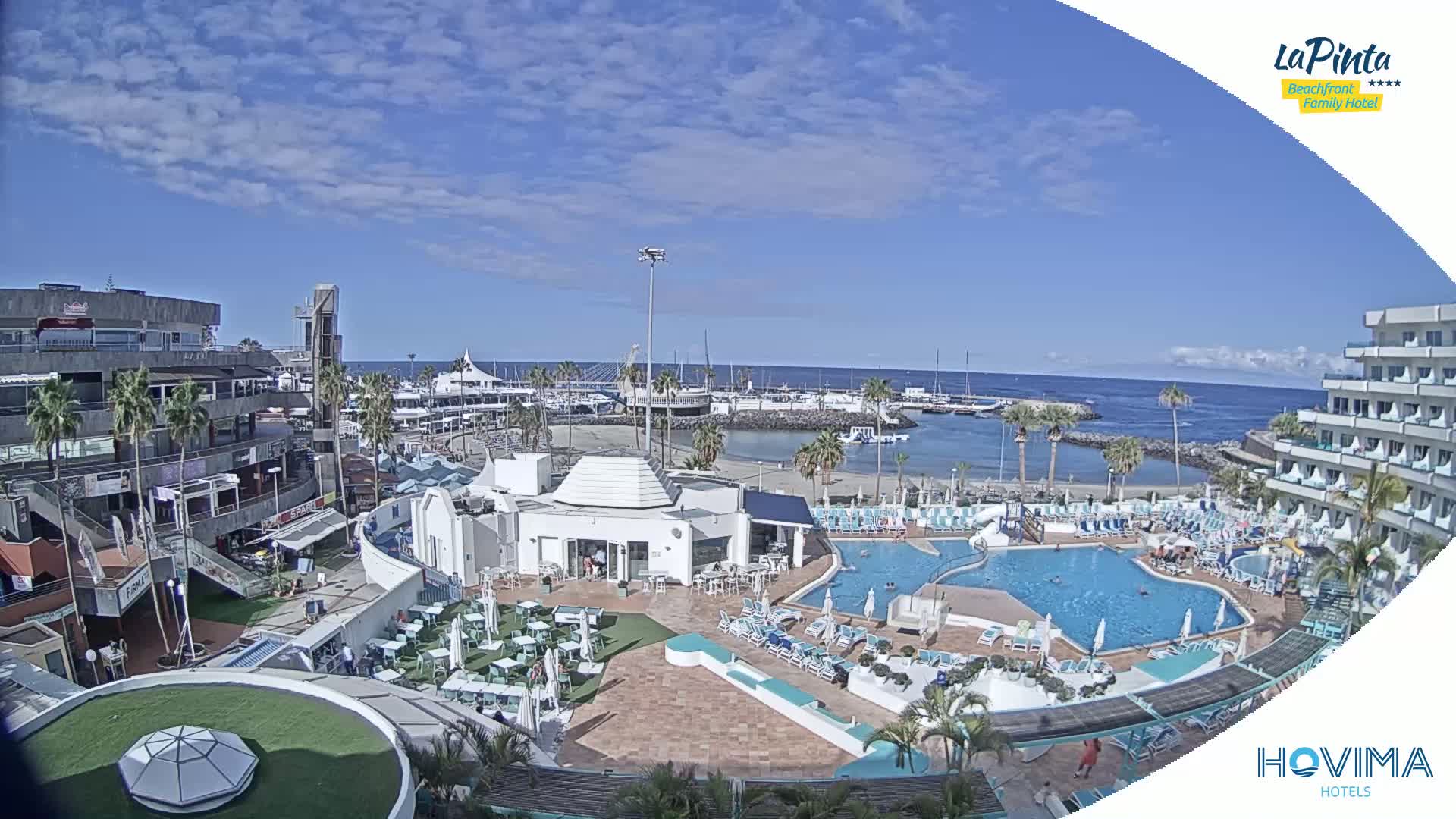 An aerial view shows a resort with a swimming pool and a small beach next to a marina on a sunny day.