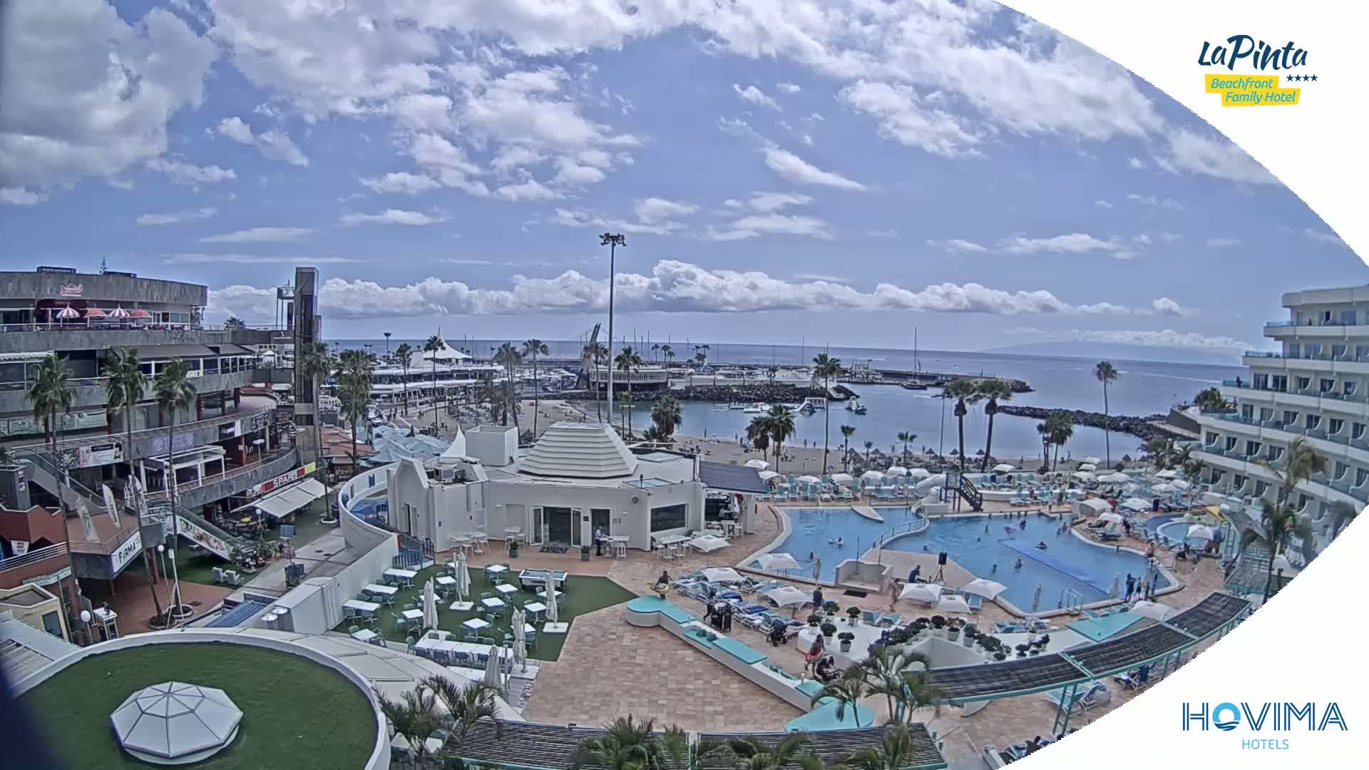 A partly cloudy day overlooking a resort pool area, beach, marina, and buildings.