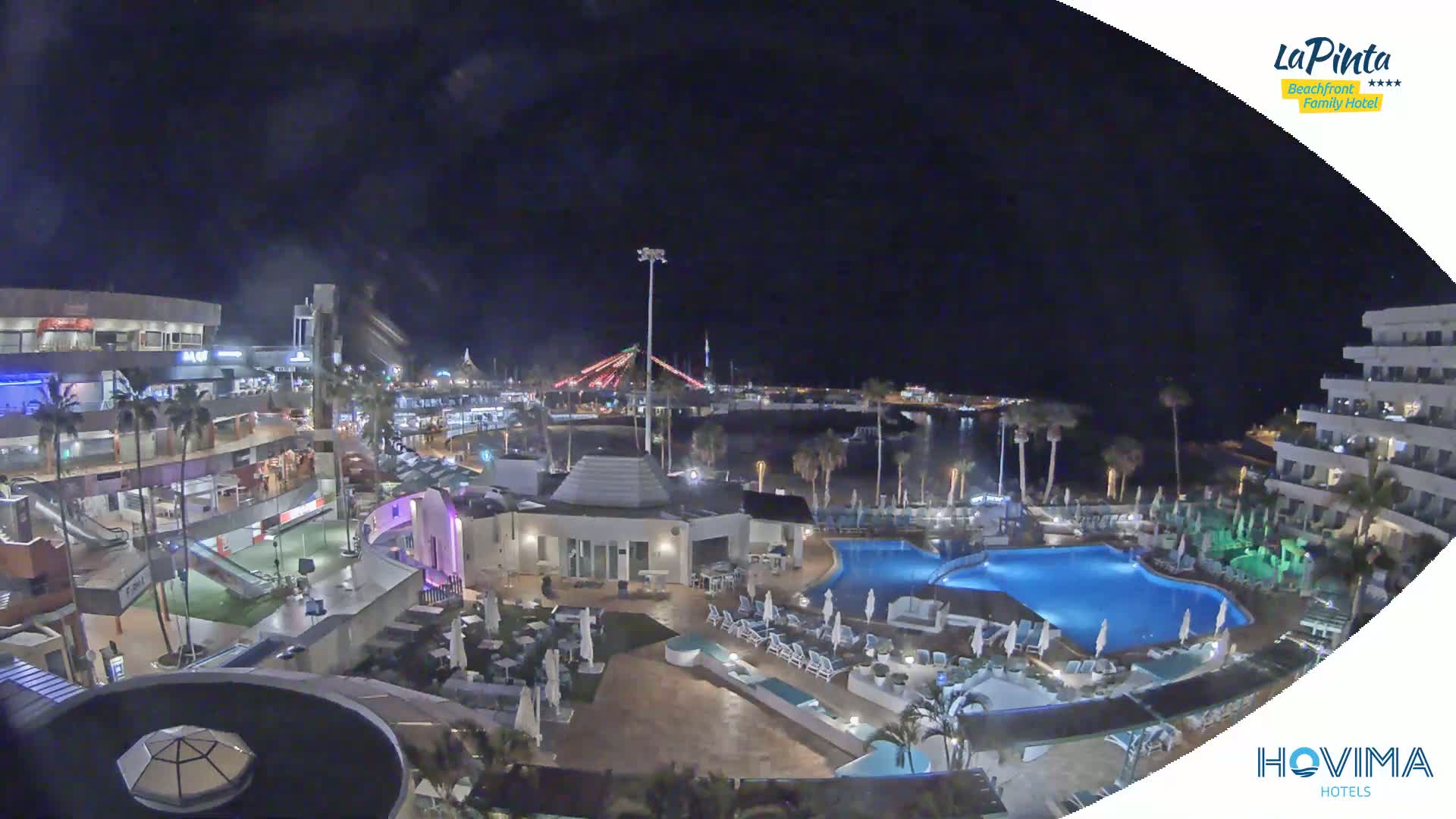 A nighttime aerial view of a resort complex with pools, surrounding buildings, and a waterfront area beyond.