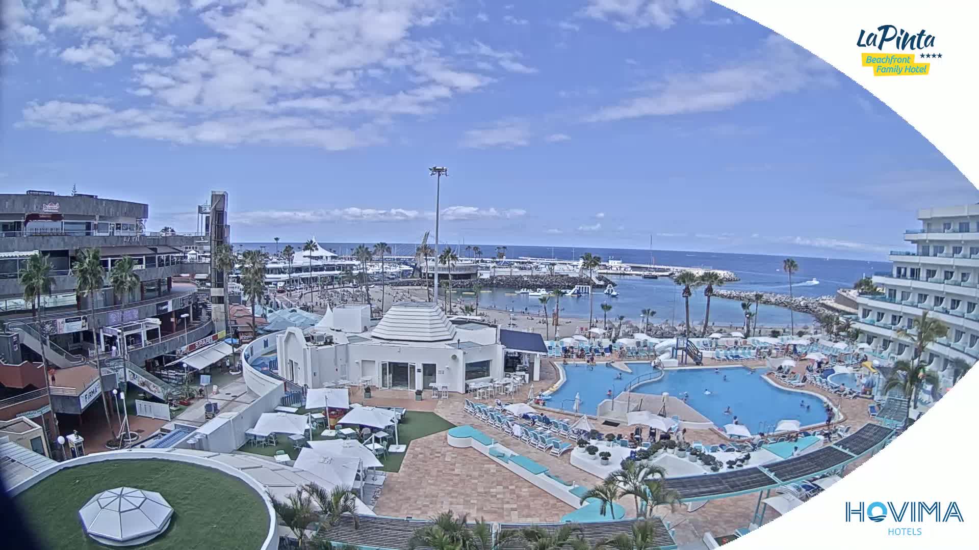A resort area with swimming pools and a beach, situated next to a harbor on a sunny day with partly cloudy skies.