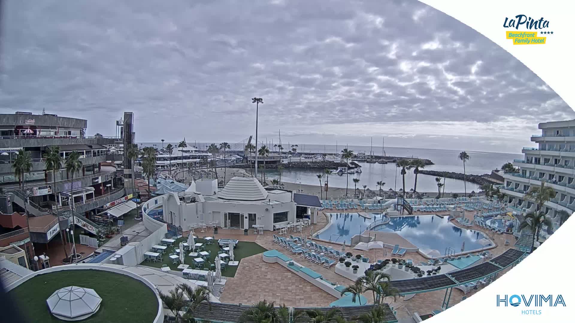An overcast day reveals a resort area with a marina, a beach, multiple pools, and buildings, including shops and hotels.