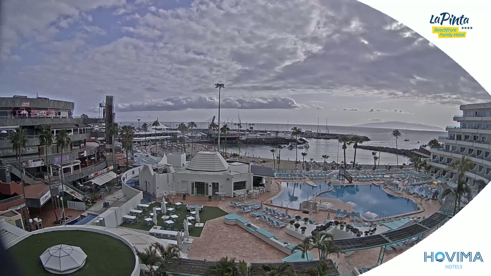 An outdoor view of a resort complex with a swimming pool, beach, marina, and shops under a partly cloudy sky.