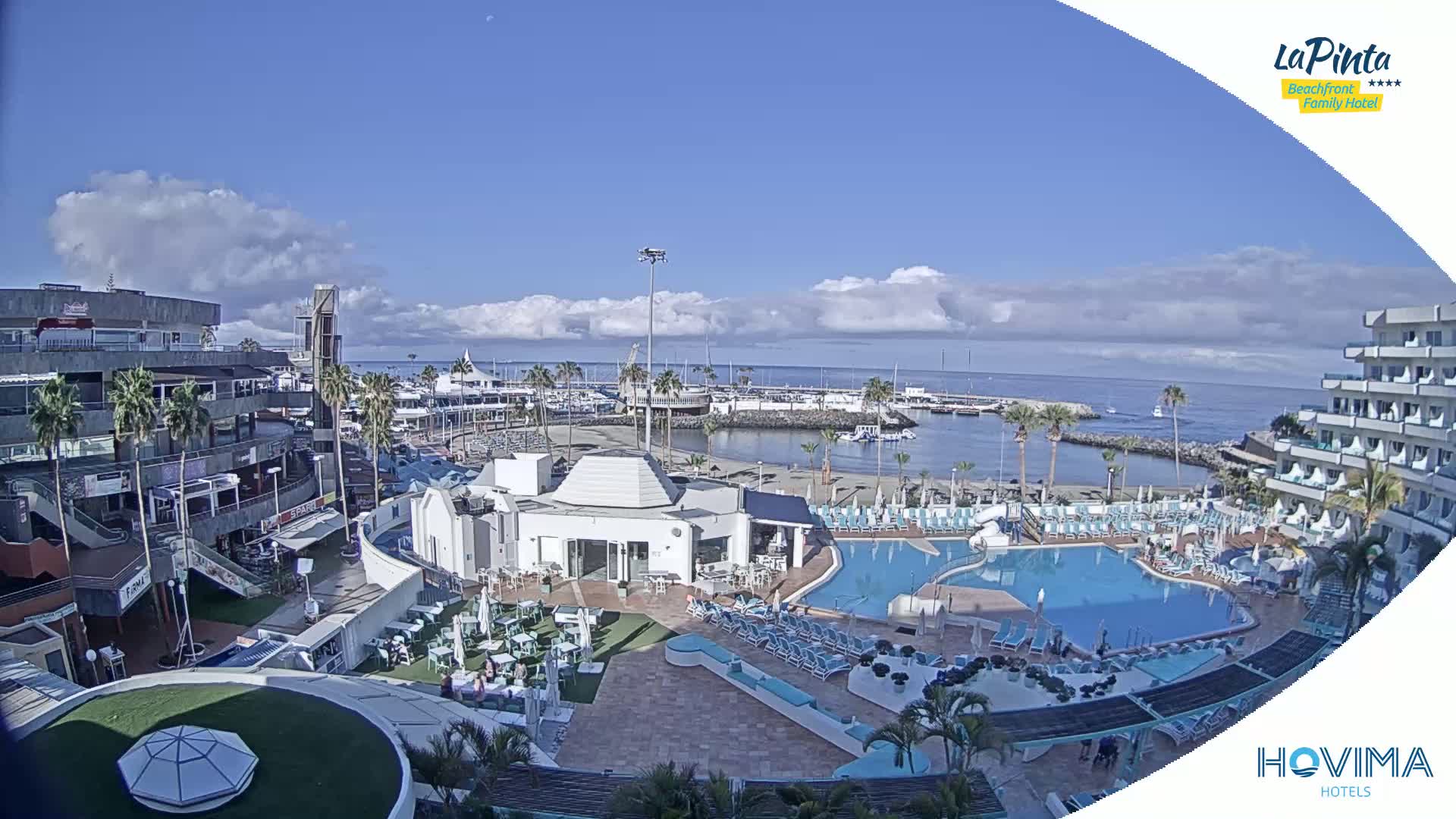 A resort with a swimming pool overlooks a marina and calm ocean under a partly cloudy sky.