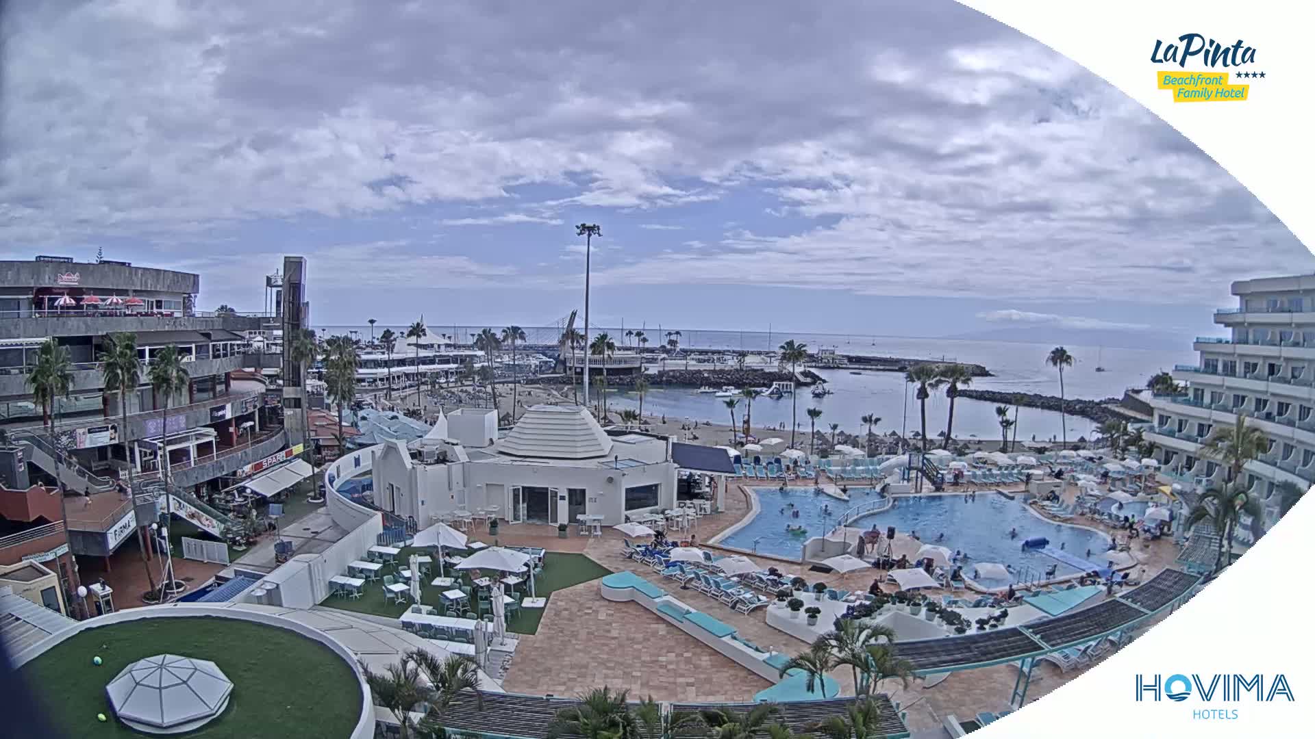 An aerial view shows a resort with a swimming pool, beach, and shops under a partly cloudy sky.