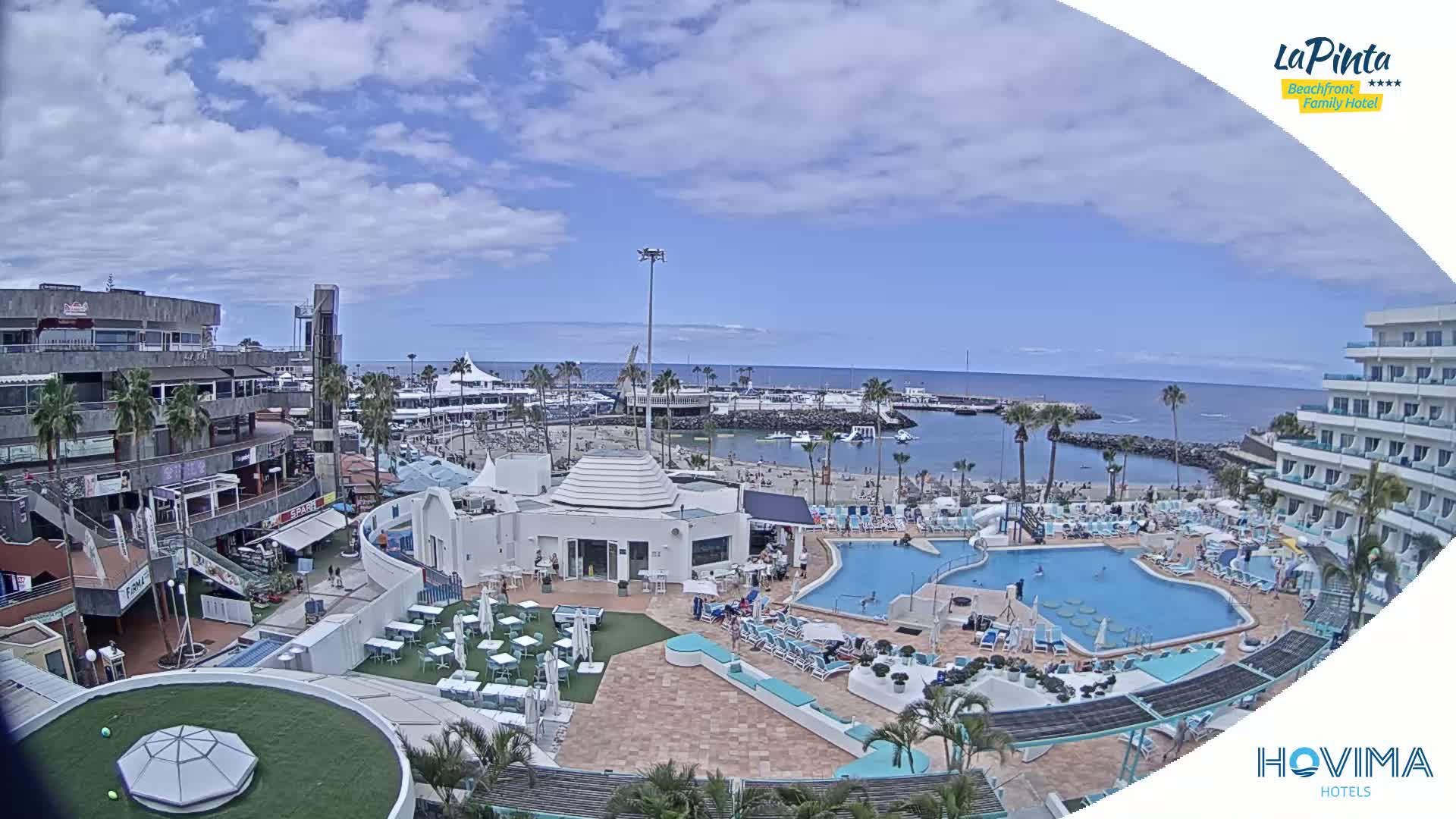 A sunny day overlooking a resort pool area, a marina, and a sandy beach with a calm ocean in the background.