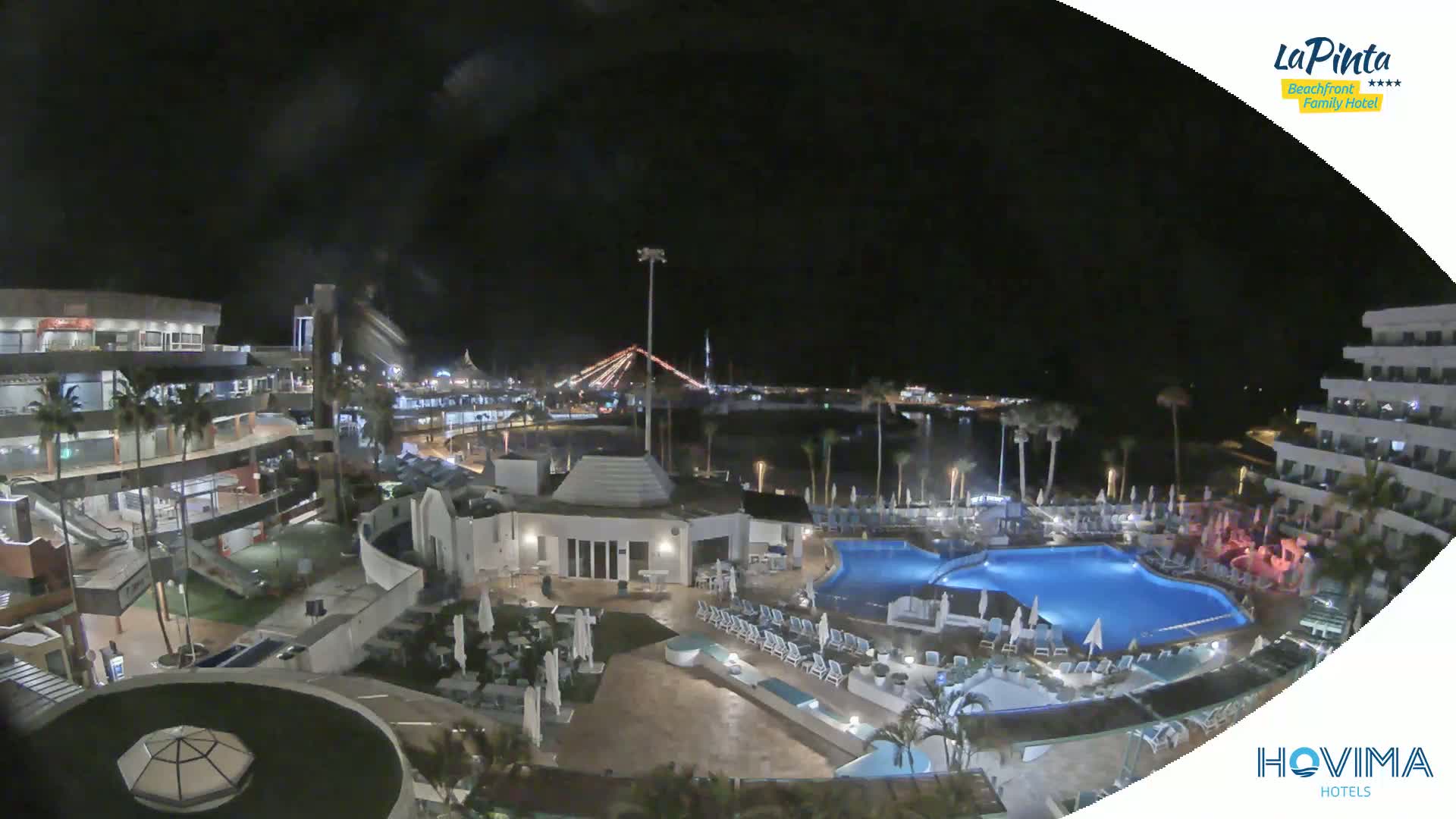 A nighttime aerial view of a resort complex with swimming pools and palm trees, illuminated by artificial lights under a clear, dark sky.