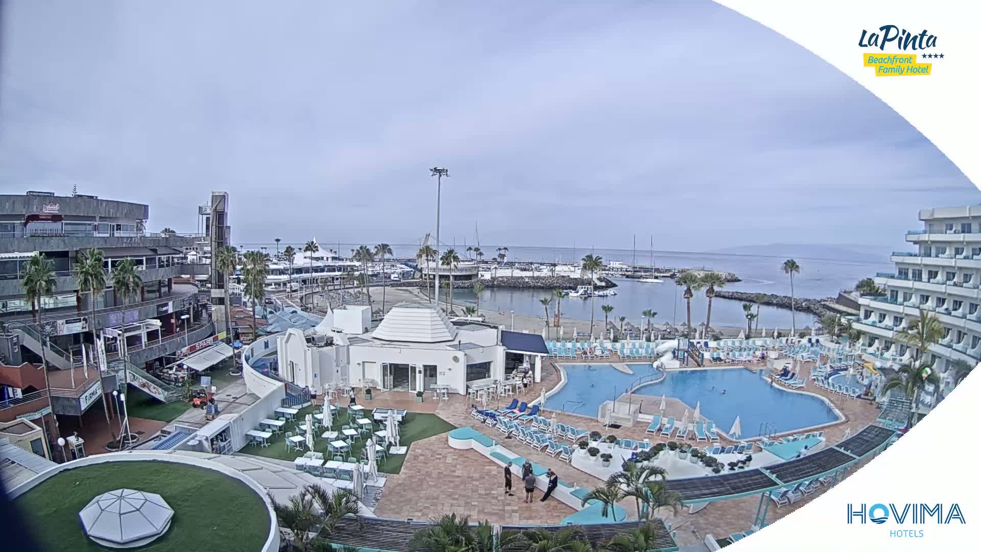 An overcast day shows a resort with a swimming pool overlooking a marina and calm ocean.