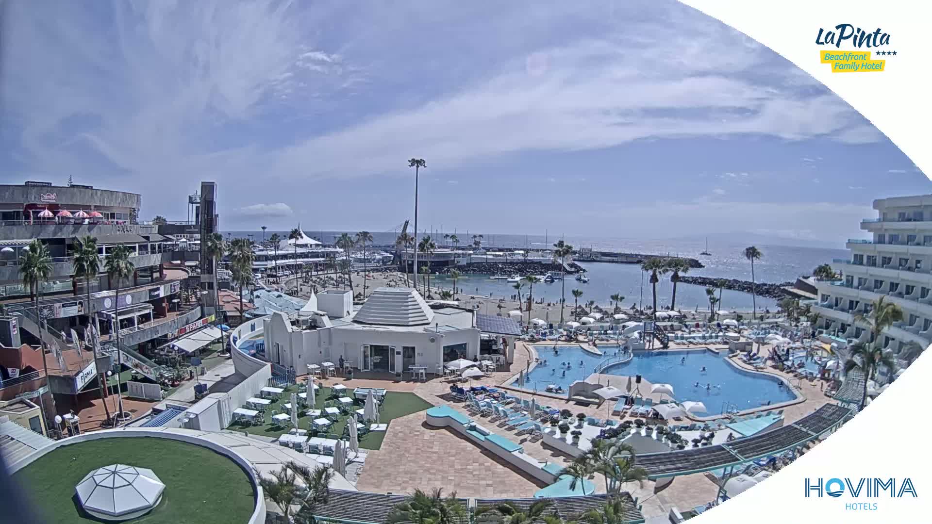 An aerial view shows a resort area with swimming pools, a beach, and shops under a sunny sky.
